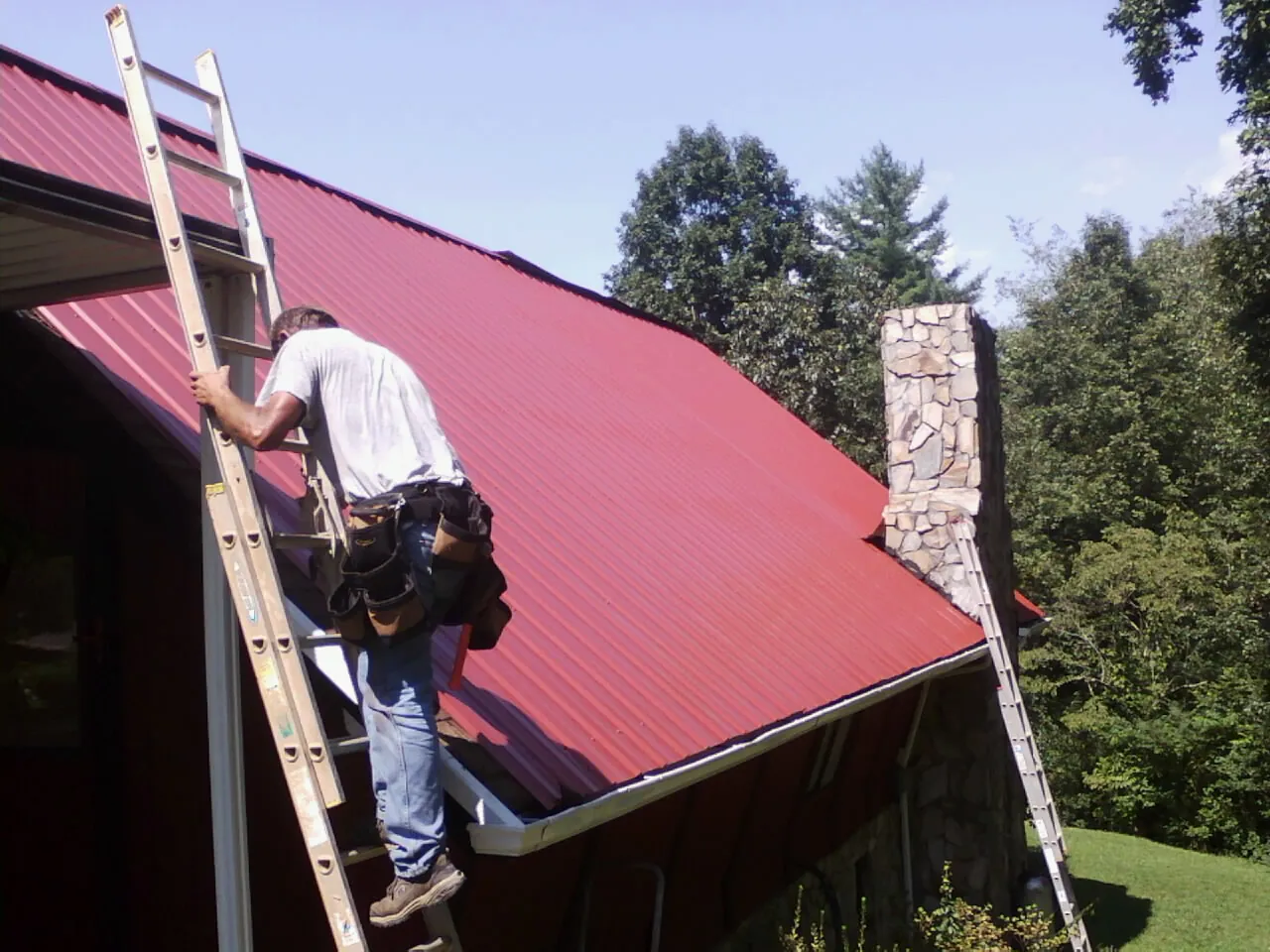 Roofer on a ladder beside a red metal roof and stone chimney — Soffit & Fascia Repair by Roofing Brothers in Saint Helena